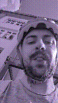a selfie of a white person, wearing either headphones or a helmet, making an odd expression. the photo is edited to be colorized into a monochrome purple-ish color palette.