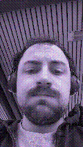a selfie of a white person, wearing either headphones or a helmet, making an odd expression. the photo is edited to be colorized into a monochrome purple-ish color palette.