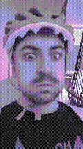 a selfie of a white person, wearing either headphones or a helmet, making an odd expression. the photo is edited to be colorized into a monochrome purple-ish color palette.