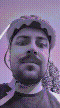 a selfie of a white person, wearing either headphones or a helmet, making an odd expression. the photo is edited to be colorized into a monochrome purple-ish color palette.