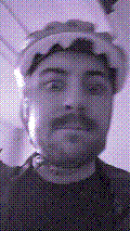 a selfie of a white person, wearing either headphones or a helmet, making an odd expression. the photo is edited to be colorized into a monochrome purple-ish color palette.